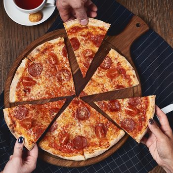 Pizza slices, being grabbed by hands, rest on a round wooden board; nearby, a cup of tea, a cookie, phone, and salt shaker.