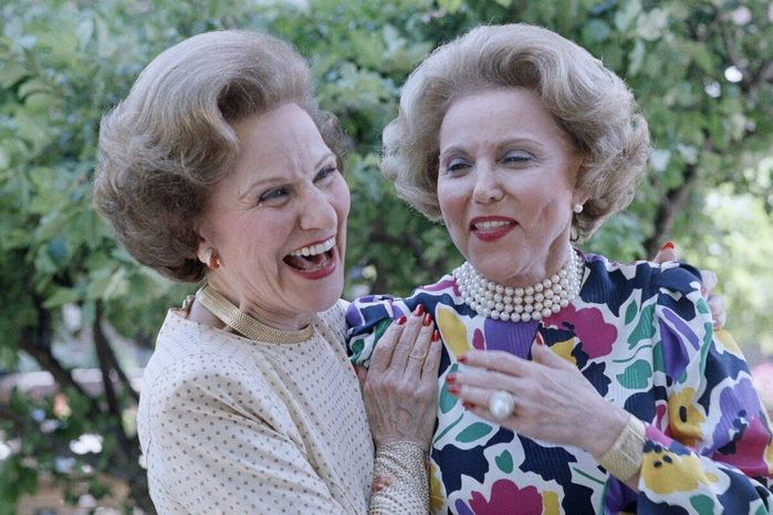Two women laughing, one in a polka-dot blouse, the other in a colorful floral dress with pearls, standing outdoors among green trees.