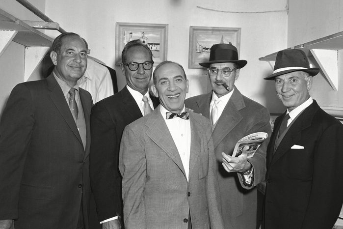 Five men stand smiling in suits and hats, one holding a magazine, inside a room with paintings on the walls.