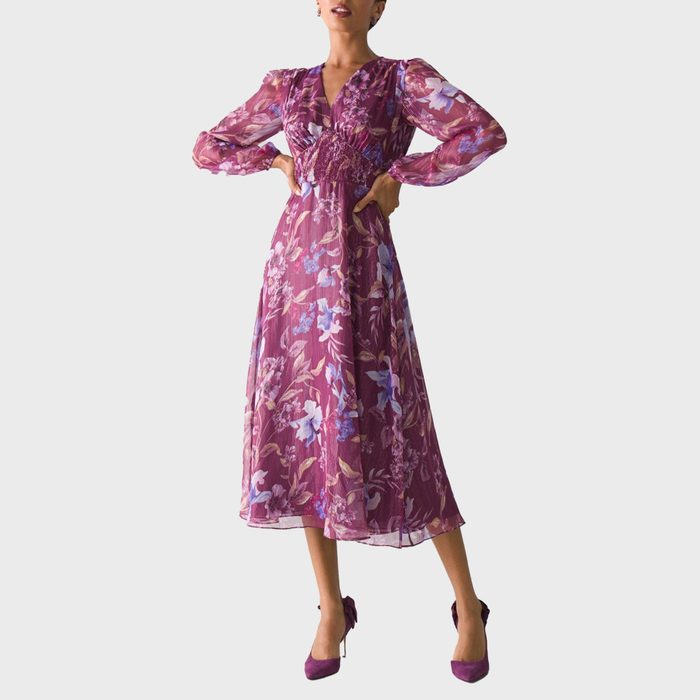 A person poses confidently wearing a long, floral purple dress with puffed sleeves, set against a plain white background.