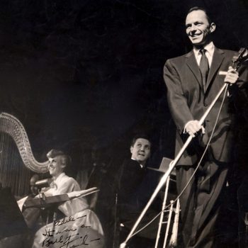 Man holding microphone smiles on stage; musicians play behind him with harp and piano; autograph reads "Adele, Here we are 'Budd' Talk."