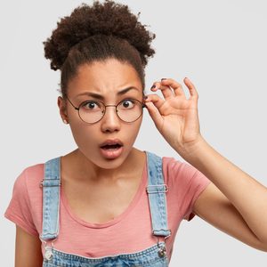 Person adjusts round glasses, expression surprised, wearing pink shirt and denim overalls, against plain gray background.