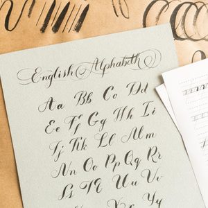 Calligraphy sheet displays cursive English alphabet. Next to the sheet, two brushes rest on brown paper with practice strokes.