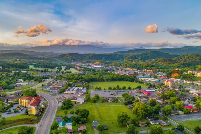 Aerial view shows a small town amidst lush greenery and distant mountains under a clear sky with scattered clouds.