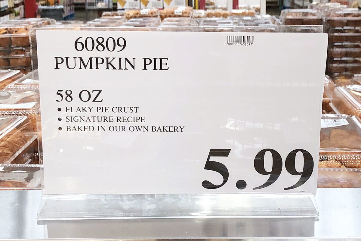 Sign displays "Pumpkin Pie," 58 oz, $5.99. Pies are stacked in transparent containers on metal tables in a warehouse-style store.
