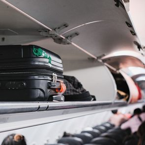 Suitcase rests inside an airplane's open overhead storage compartment, with passengers seated below in a dimly lit cabin.
