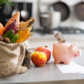 A piggy bank with cash stands near apples and groceries on a kitchen counter.