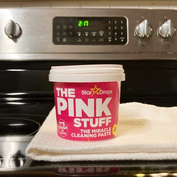 Container of "The Pink Stuff" cleaning paste rests on a cloth, set against a kitchen stove backdrop.