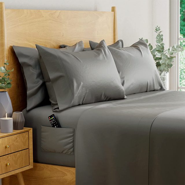 Gray bedding with a remote in pocket lies on a wooden bed, beside a nightstand with candles and eucalyptus in a vase.