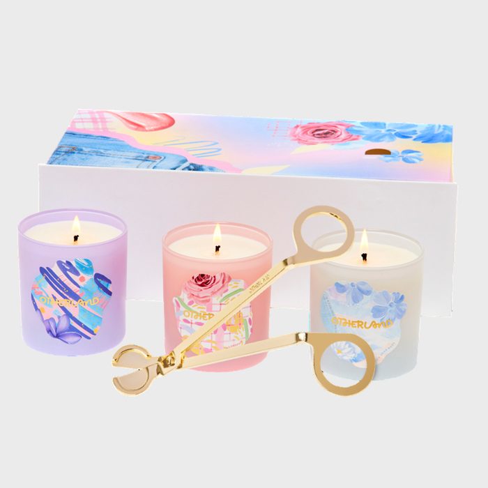 Three lit candles displayed, seen with a wick trimmer, are set against a colorful, floral-themed box background. Text: 