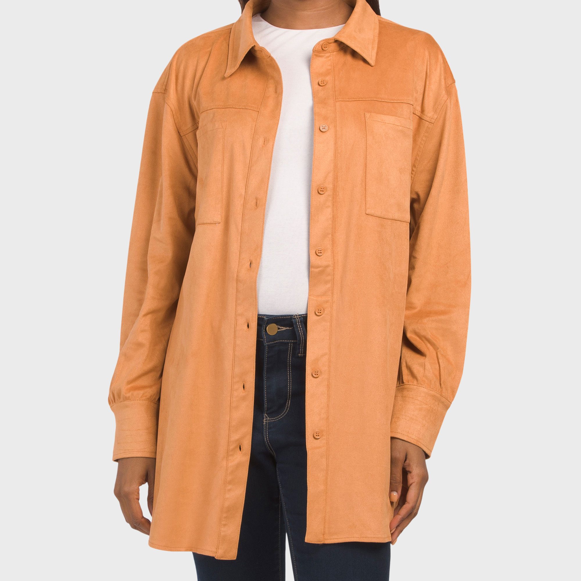 A person wears a tan button-up suede shirt over a white top and dark jeans, standing against a plain background.