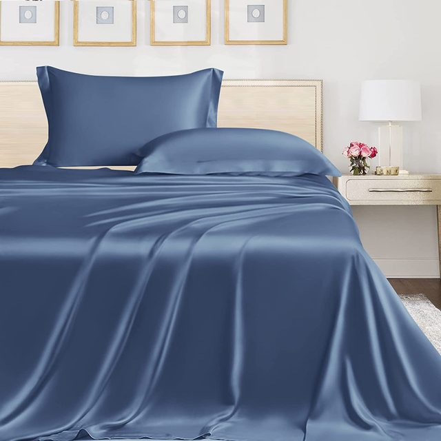 Blue satin bedding lies smoothly on a bed with wooden headboard, beside a nightstand with a lamp and flowers, in a minimalist bedroom.