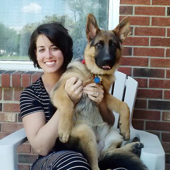 A woman holds a German Shepherd puppy, sitting on a chair against a brick wall with a window.