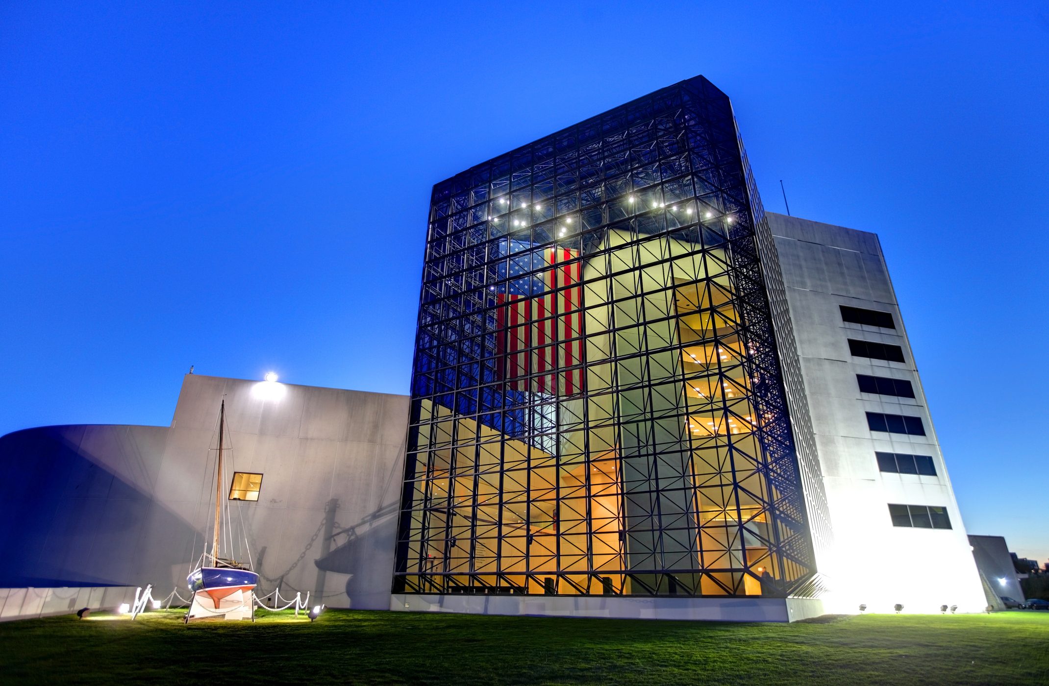 Glass building reflects lights, showcasing an American flag inside. Small sailboat is displayed outside on green grass under a clear blue evening sky.