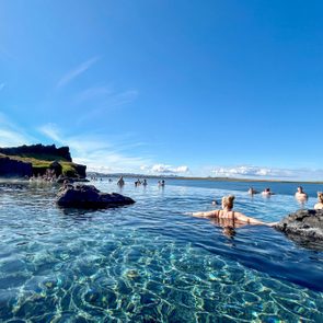 People relax in a clear, tranquil hot spring, surrounded by rocky formations and blue skies.
