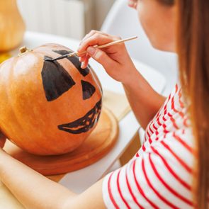 Pumpkin being painted with a black jack-o'-lantern face, surrounded by Halloween-themed decorations on a table.