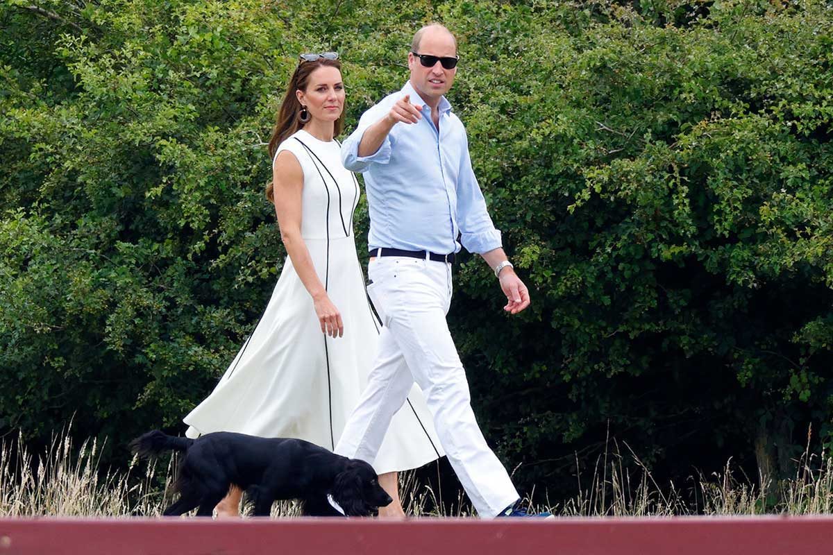 A man and woman walk a black dog on a path, pointing towards something, surrounded by dense green foliage.