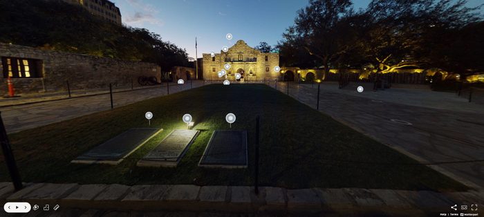 Night view of the Alamo in San Antonio, Texas, with illuminated historic plaques and gravestones in the foreground, surrounded by trees and softly lit pathways.