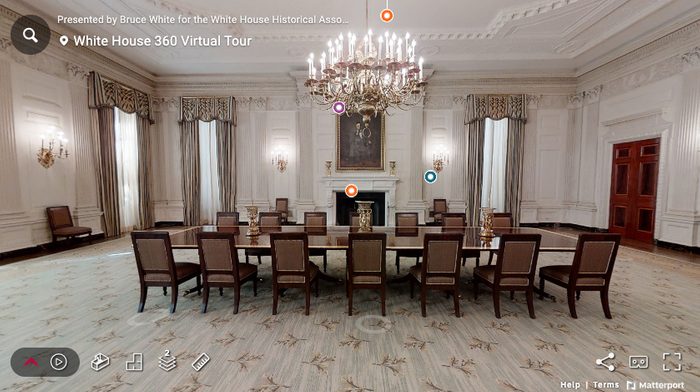 A large, elegant dining room with a long table surrounded by chairs, ornate chandeliers, tall windows with drapes, and a fireplace beneath a portrait. Virtual tour interface elements are visible on the image.